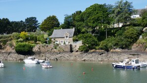 Une scène côtière avec des bateaux amarrés dans un port, un bâtiment en pierre doté d’un toit en pente, et une végétation luxuriante en arrière-plan.