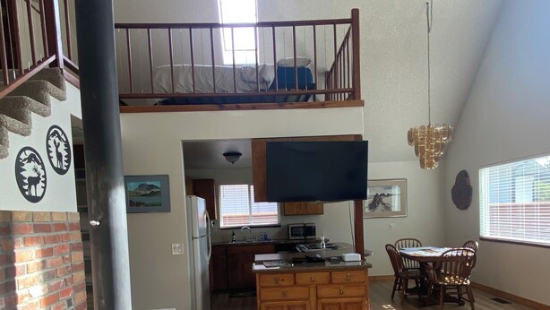 A spacious living area with a high ceiling, a fireplace, a dining table with chairs, and a kitchen in the background.