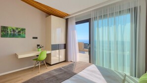 A modern hotel room with a bed, a desk with a chair, a large window with sheer curtains, and a view of the sea. - Lovely home in Smrika (Smrika)