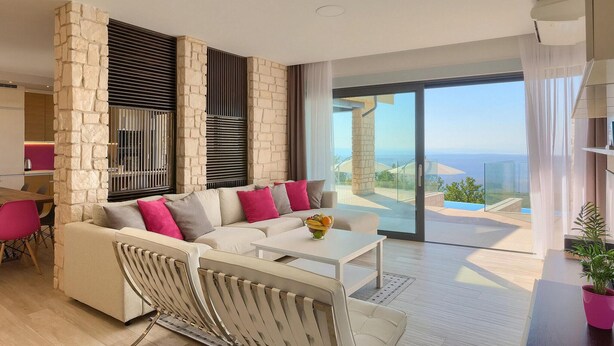 A modern living room with a stone accent wall, a sofa with colorful cushions, a white coffee table, and a view of the sea through large glass doors. - Lovely home in Smrika (Smrika)