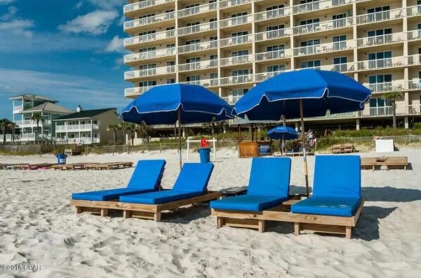 Ein Strand mit blauen Liegestühlen und Sonnenschirmen, im Hintergrund ein Hotelgebäude und ein klarer Himmel.