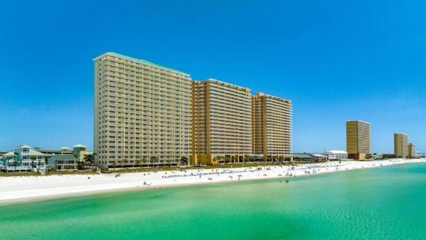 Un hotel frente a la playa con varios edificios altos, una playa de arena y aguas turquesas cristalinas.