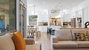 Uma cozinha moderna com armários brancos, uma sala de jantar com mesa e cadeiras, e uma sala de estar com sofá e almofadas.
