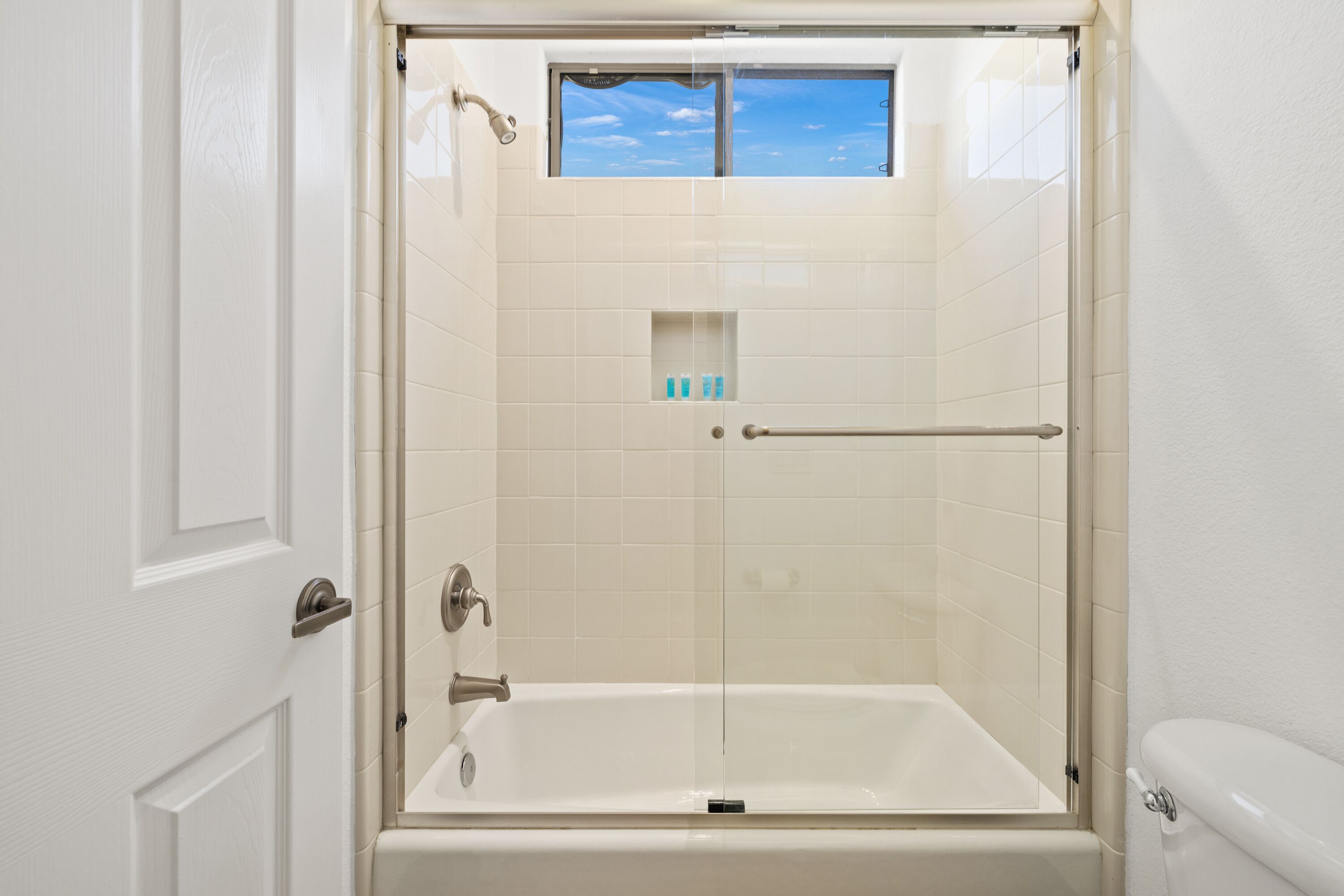 Une douche à l’italienne avec une porte en verre, une fenêtre au-dessus de la douche et une petite étagère pour les articles de toilette.