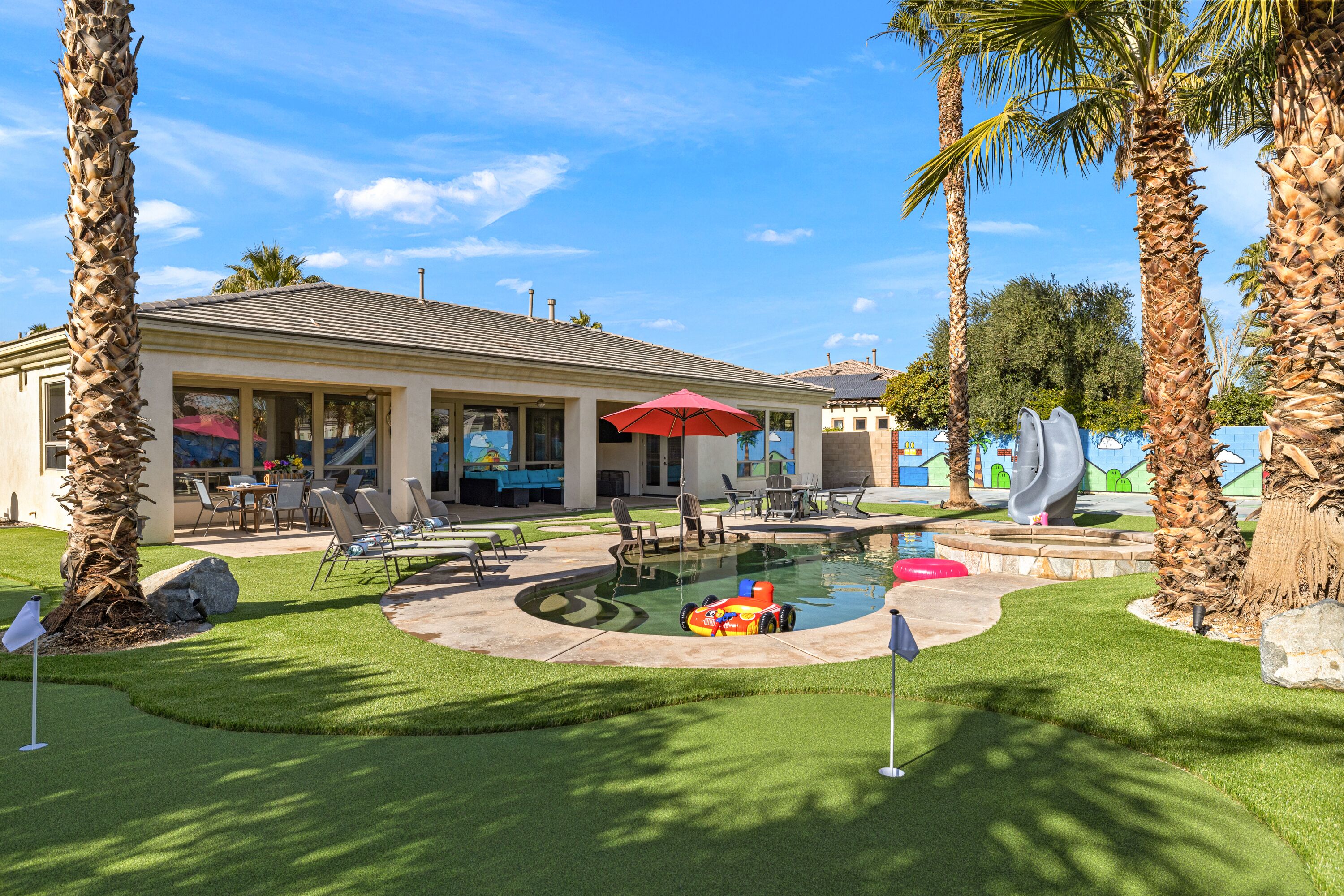 A pool area with a lounge chair, umbrella, and palm trees.