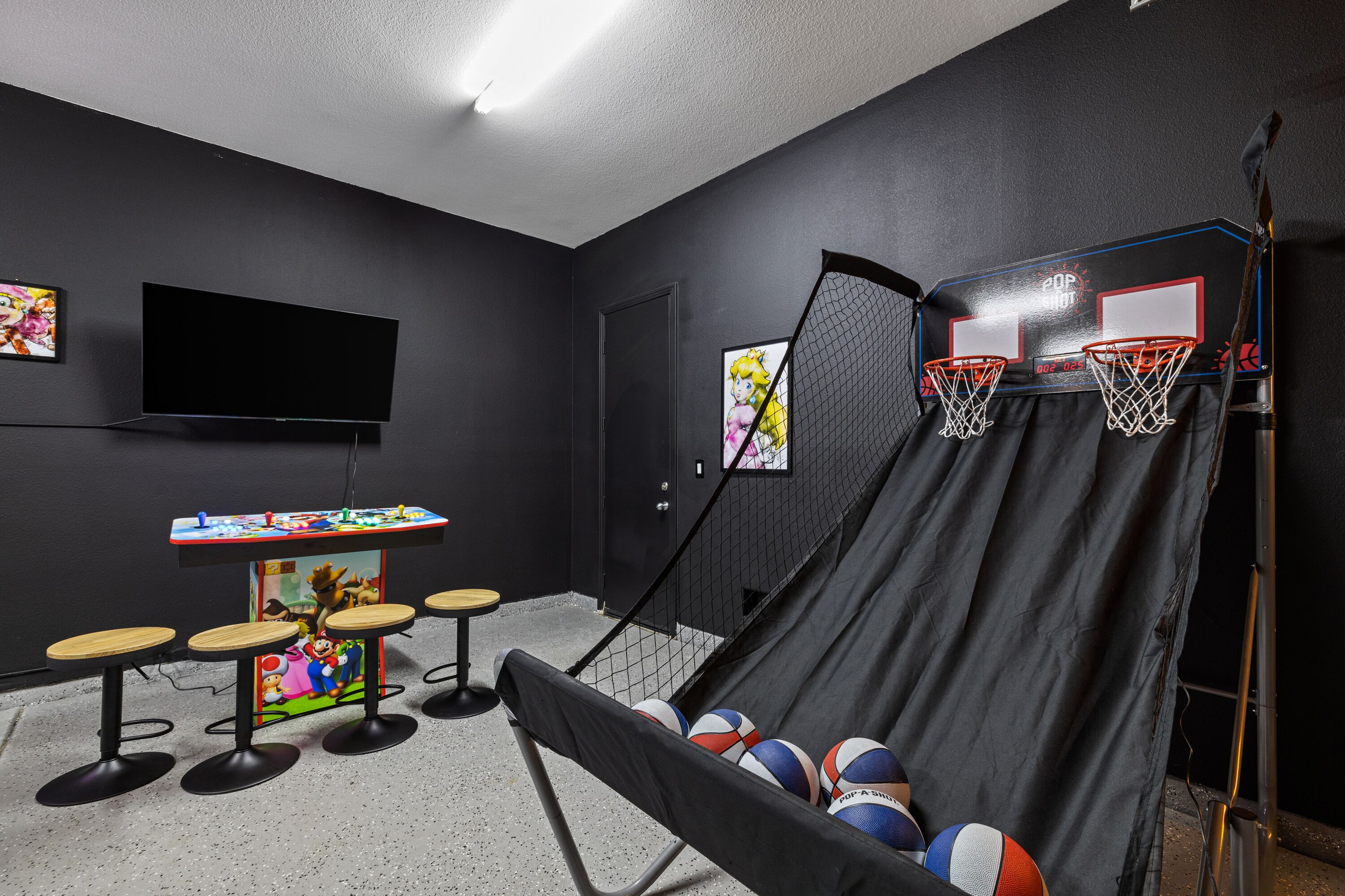Une pièce avec un mur noir, un téléviseur à écran plat, une table décorée sur le thème de Mario, quatre tabourets, un panier de basket et un toboggan.
