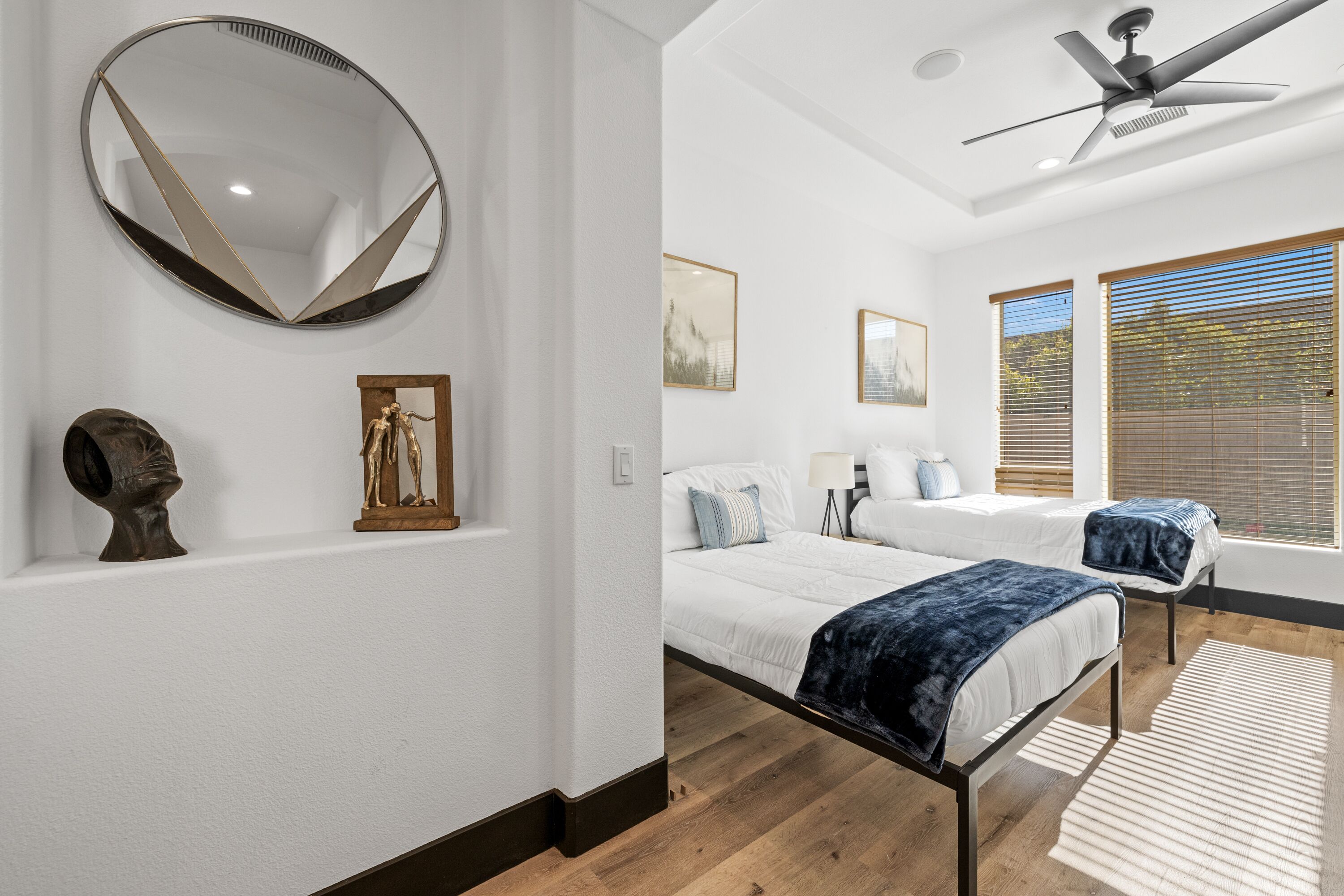 A modern bedroom with a large round mirror, two beds, a ceiling fan, and wooden flooring.