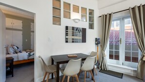 A compact room with a bed, a dining table with chairs, a wall-mounted light fixture, and a window with curtains. - The Nest - Central, Sunny & Spacious Loft (Te Aro)