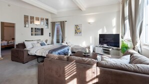 A well-lit living room with a large sofa, a bed, a TV, and a window with curtains. - The Nest - Central, Sunny & Spacious Loft (Te Aro)