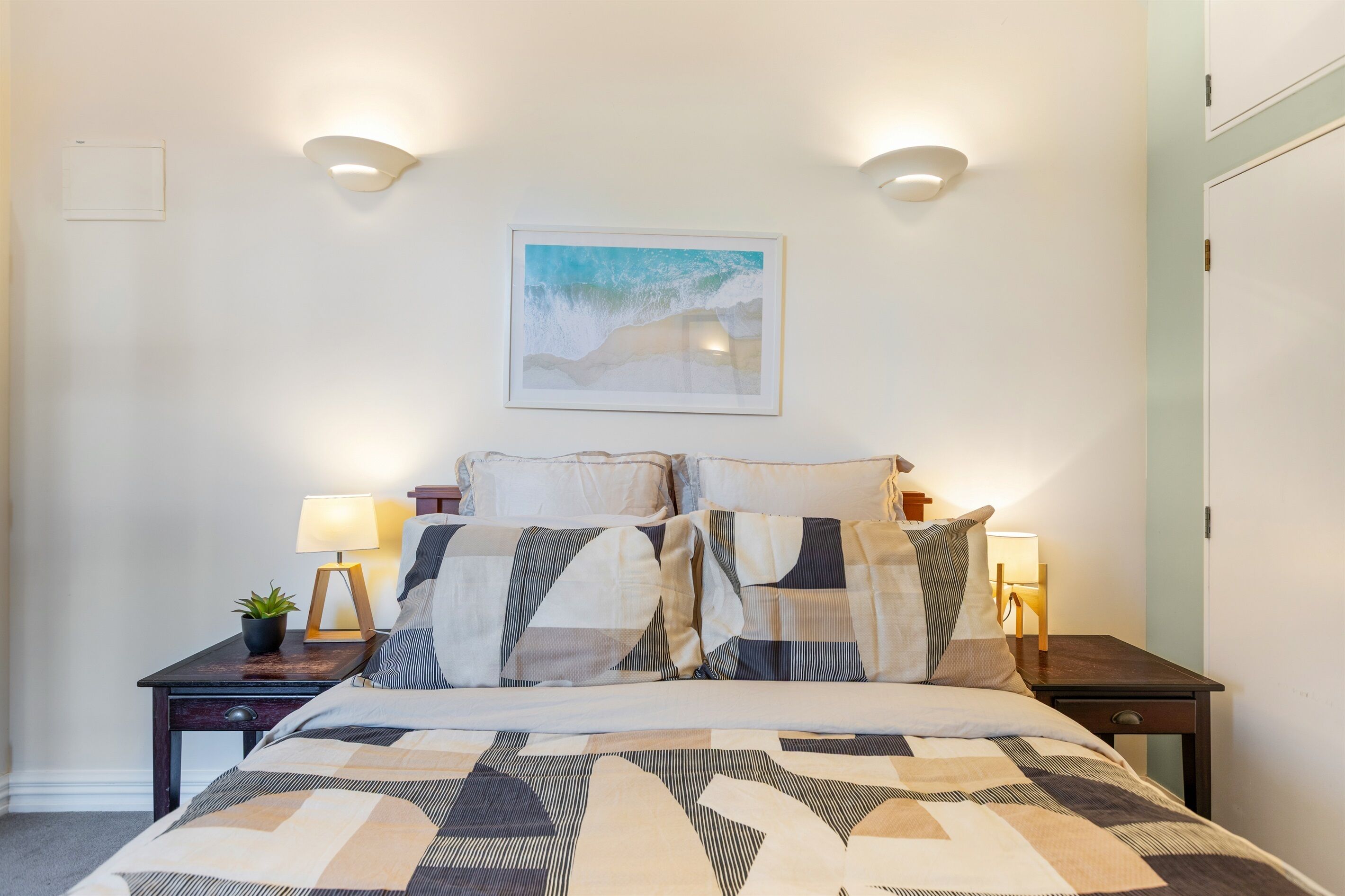 A neatly made bed with patterned bedding, two bedside tables with lamps, a framed picture on the wall, and a door to the right.