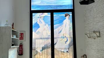 A room with a large glass door featuring a painting of two figures in white dresses, a white brick wall, a green sofa, a small table, and a ceiling light fixture.