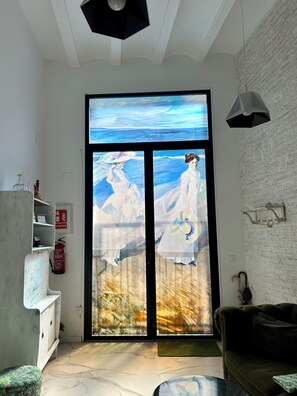 A room with a large glass door featuring a painting of two figures in white dresses, a white brick wall, a green sofa, a small table, and a ceiling light fixture. - Loft con Mucha luz en el Centro. Barrio Ruzafa (València)