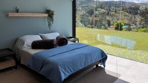 A modern bedroom with a large bed, a nightstand, a wooden shelf with a plant, and a view of a green landscape through a glass door. - Beautiful Finca with Jacuzzi. Large green areas, surrounded by vegetation. (Rionegro)