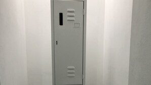 A tall, narrow metal locker with multiple compartments, standing in a corner with tiled flooring. - El Tip Viajero, Apartaments and Coworking  Lima Perú (San Martín de Porres)
