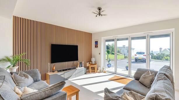 A modern living room with a flat-screen TV, grey sofas, wooden panel wall, and a view of a street through large glass doors. - 2 bedroom accommodation in Torquay (Torquay)