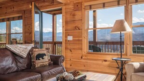 Interior de una cabaña de madera con un sofá de cuero, una mesa con objetos decorativos, una lámpara y ventanas grandes con vistas a las montañas.