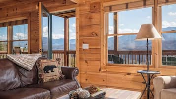 A wooden cabin interior with a leather sofa, a table with decorative items, a lamp, and large windows offering mountain views.