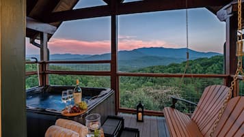 A hot tub with wine glasses and grapes, a wooden deck with a swing chair, and a scenic mountain view.