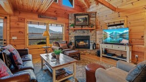 Interior de una cabaña de troncos con chimenea, televisor, sofás de cuero y vistas a montañas a través de amplios ventanales.