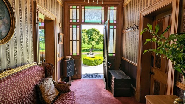A grand hallway with a chandelier, a red patterned sofa, and a view of a garden through large windows. - Mainholm Lodge (Pomahaka)