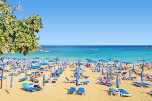 A beach with numerous sun loungers and umbrellas, a clear blue sky, and the ocean in the background.
