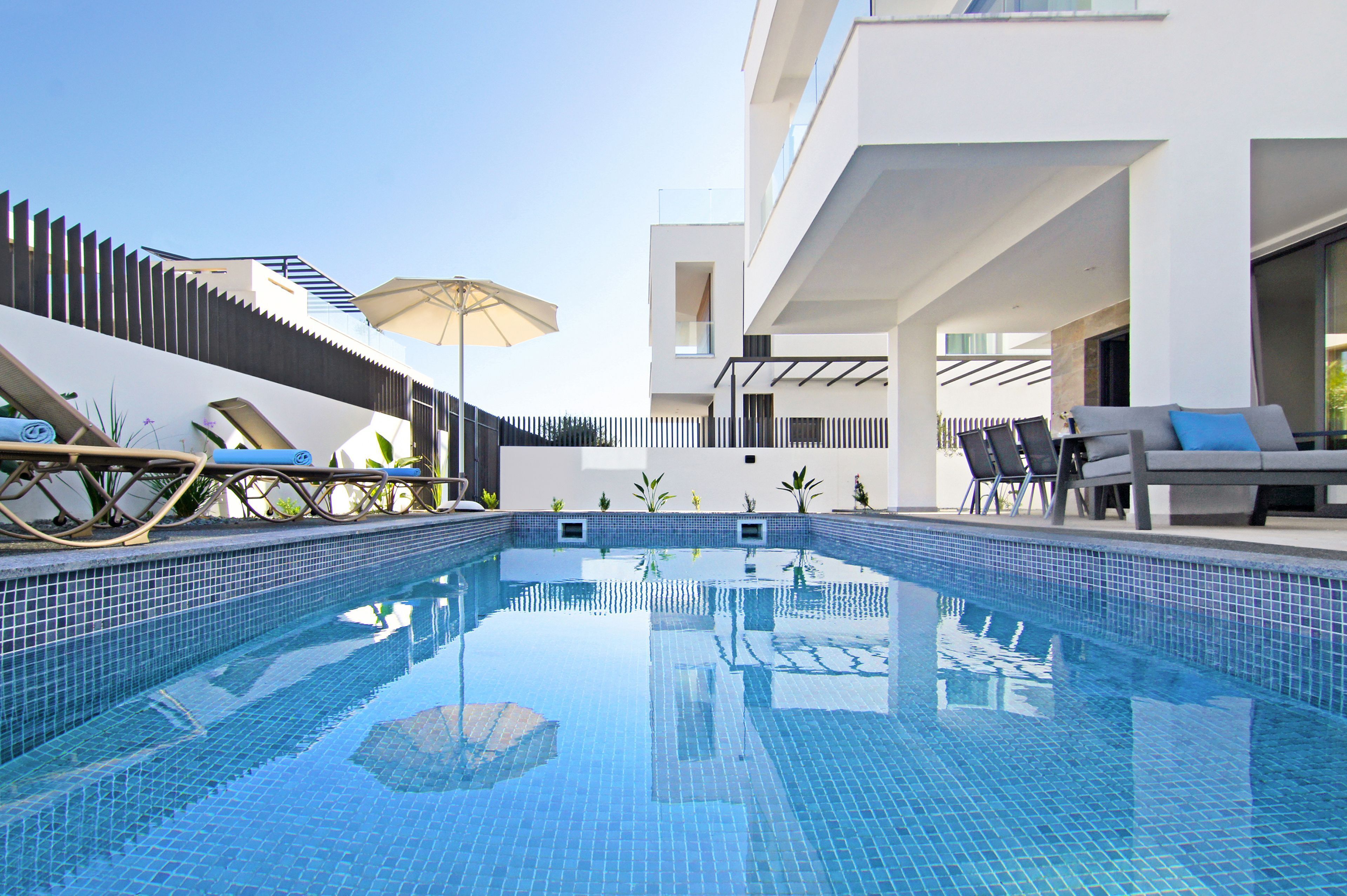 Un área de piscina moderna con sillas de descanso y sombrillas, rodeada por un edificio blanco con balcones.
