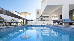 Un espace piscine moderne, doté de chaises longues et de parasols, entouré d’un bâtiment blanc agrémenté de balcons.
