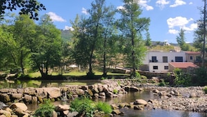 A serene riverside setting with a stone bridge, lush greenery, and a building with a red roof. - Be Alva (Oliveira do Hospital)