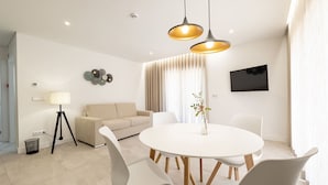 A modern living room with a round dining table, white chairs, a beige sofa, a floor lamp, and a flat-screen TV mounted on the wall. - Be Alva (Oliveira do Hospital)