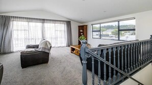 A spacious living room with a large window, a dark sofa, a wooden bookshelf, and a staircase with a black railing. - Spacious, modern, centrally located house. (Geraldine)