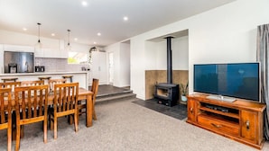 A modern living room with a TV, wooden dining table, chairs, a fireplace, and a kitchen area with white cabinets and a black stove. - Spacious, modern, centrally located house. (Geraldine)
