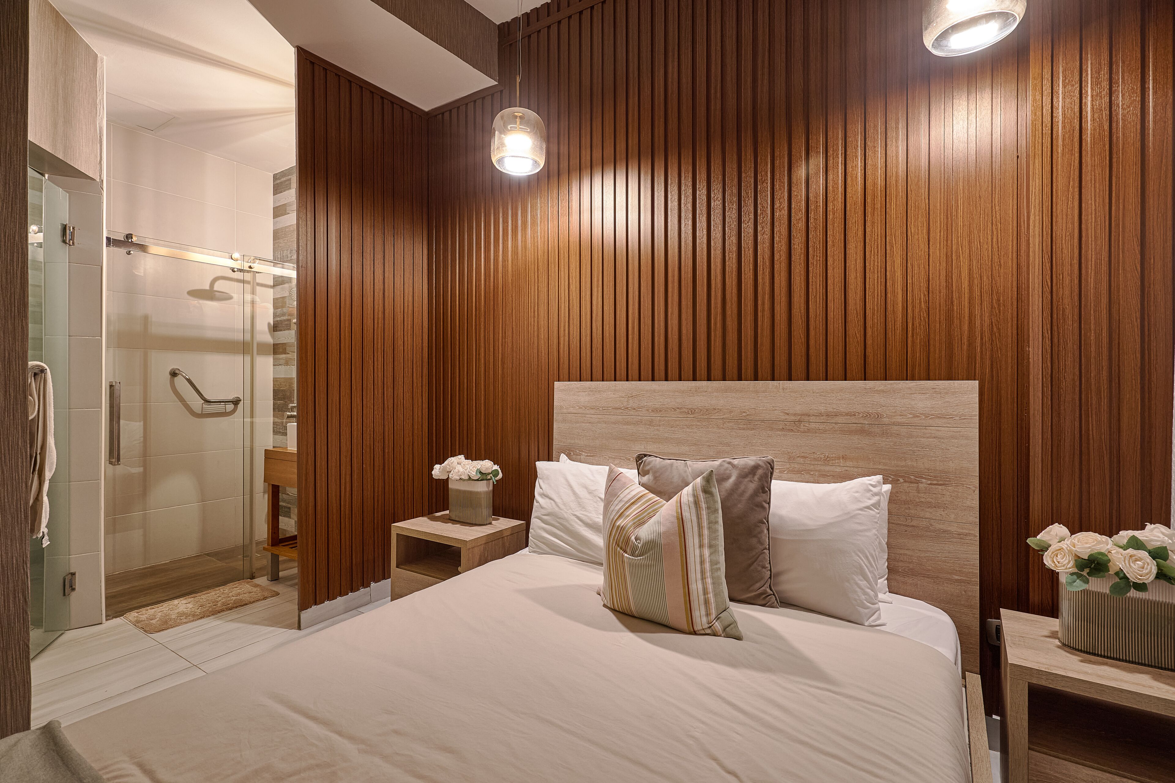 Quarto de hotel moderno com parede de painel de madeira, uma cama grande com várias almofadas, uma mesinha de cabeceira com um vaso de flores e área de chuveiro com box de vidro.