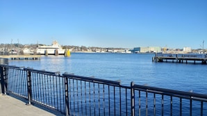 Vista al agua con un muelle, un barco grande y edificios industriales al fondo.
