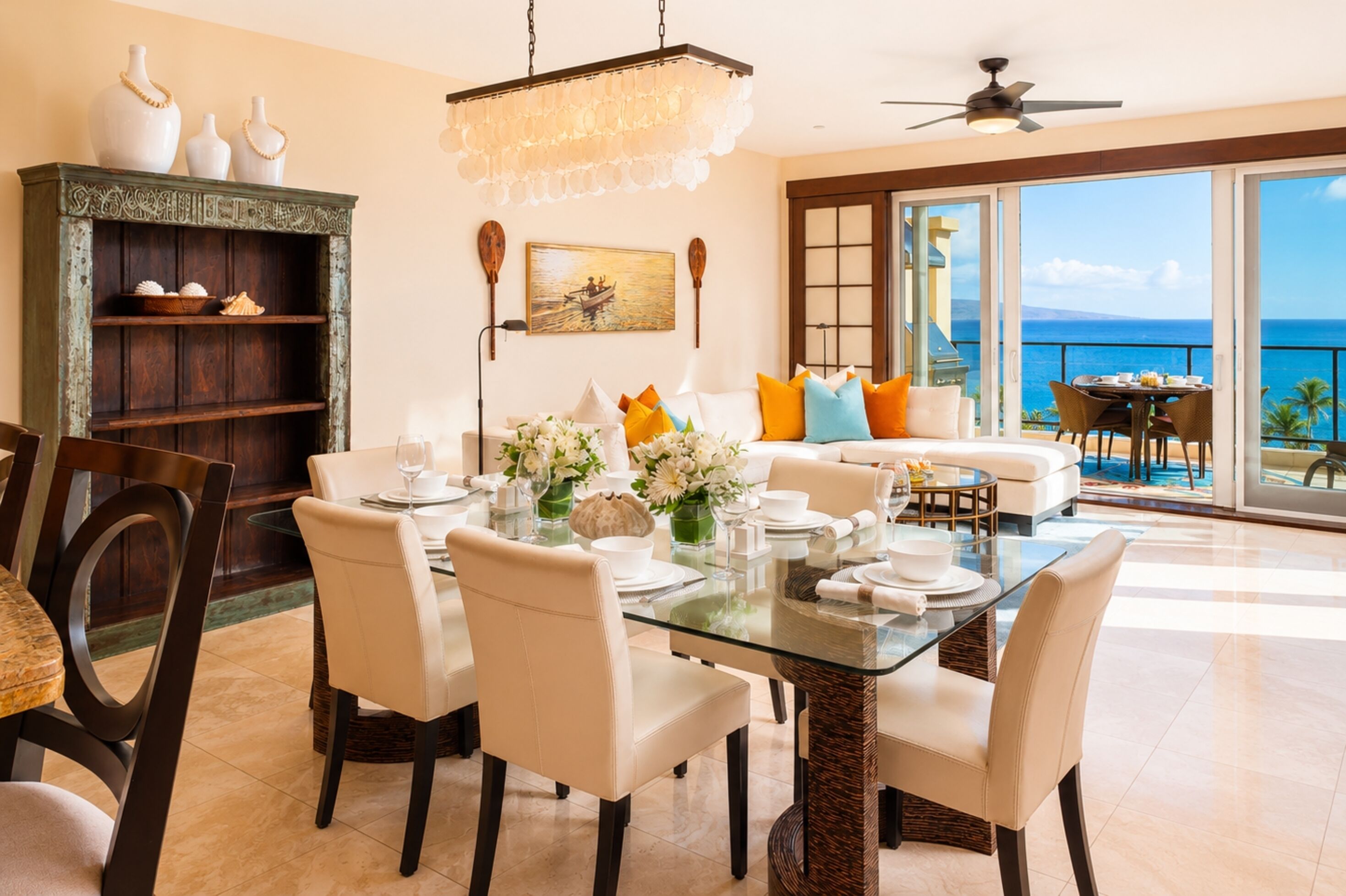 A dining area with a glass table, white chairs, and a view of the ocean through large sliding doors.