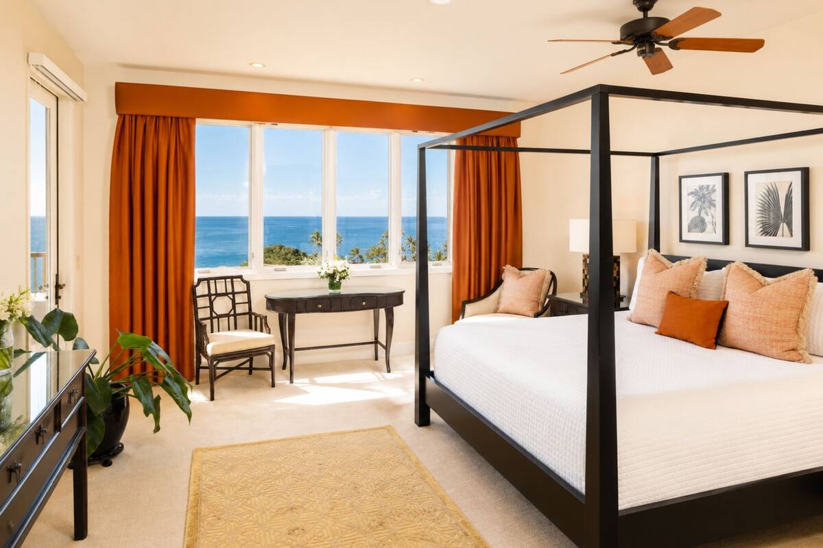 A four-poster bed with a view of the ocean, a dining table, and a chair in a room with large windows and curtains.