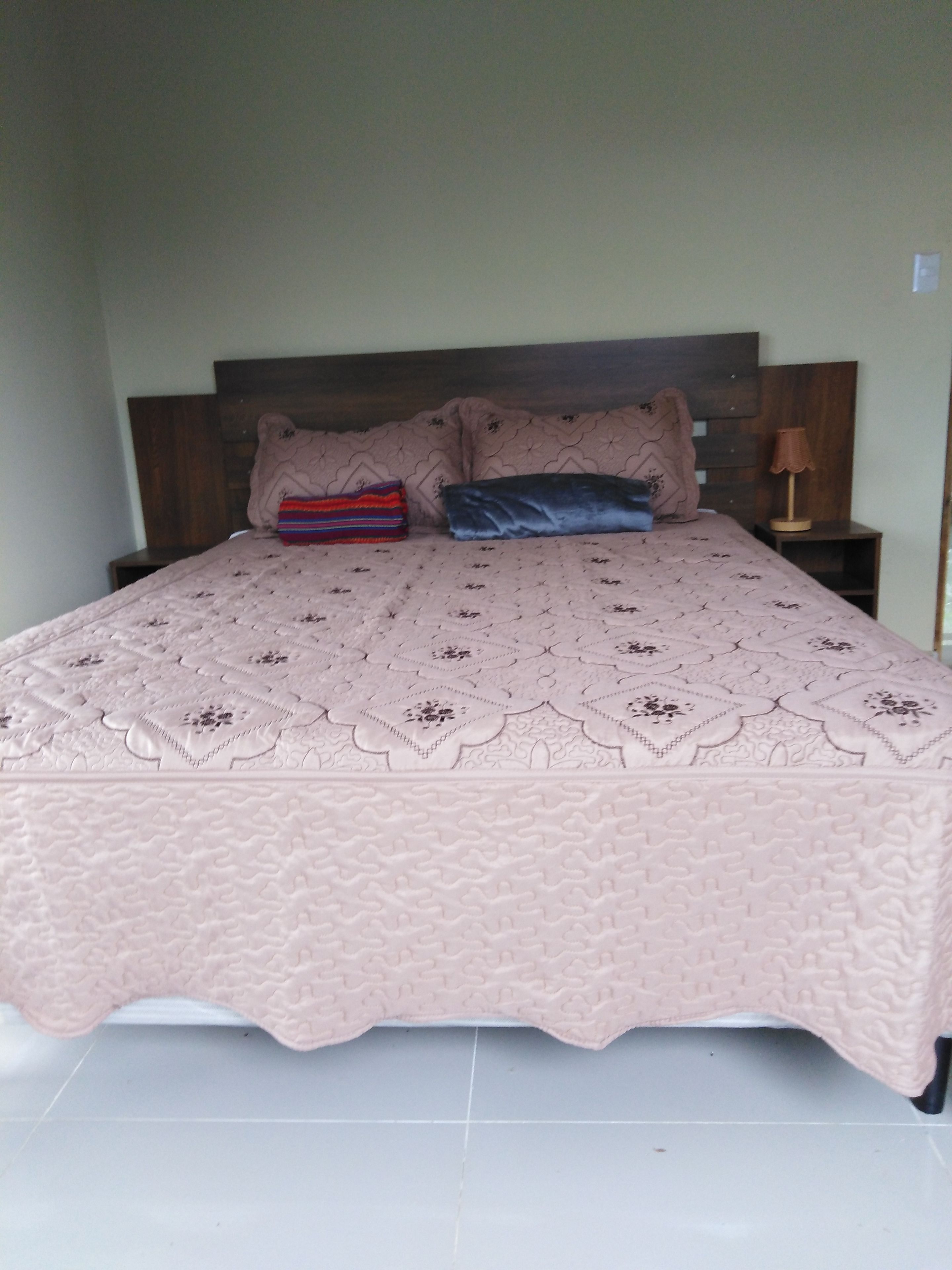 A bed with a patterned bedspread, two pillows, a bedside table with a lamp, and a wall-mounted switch.