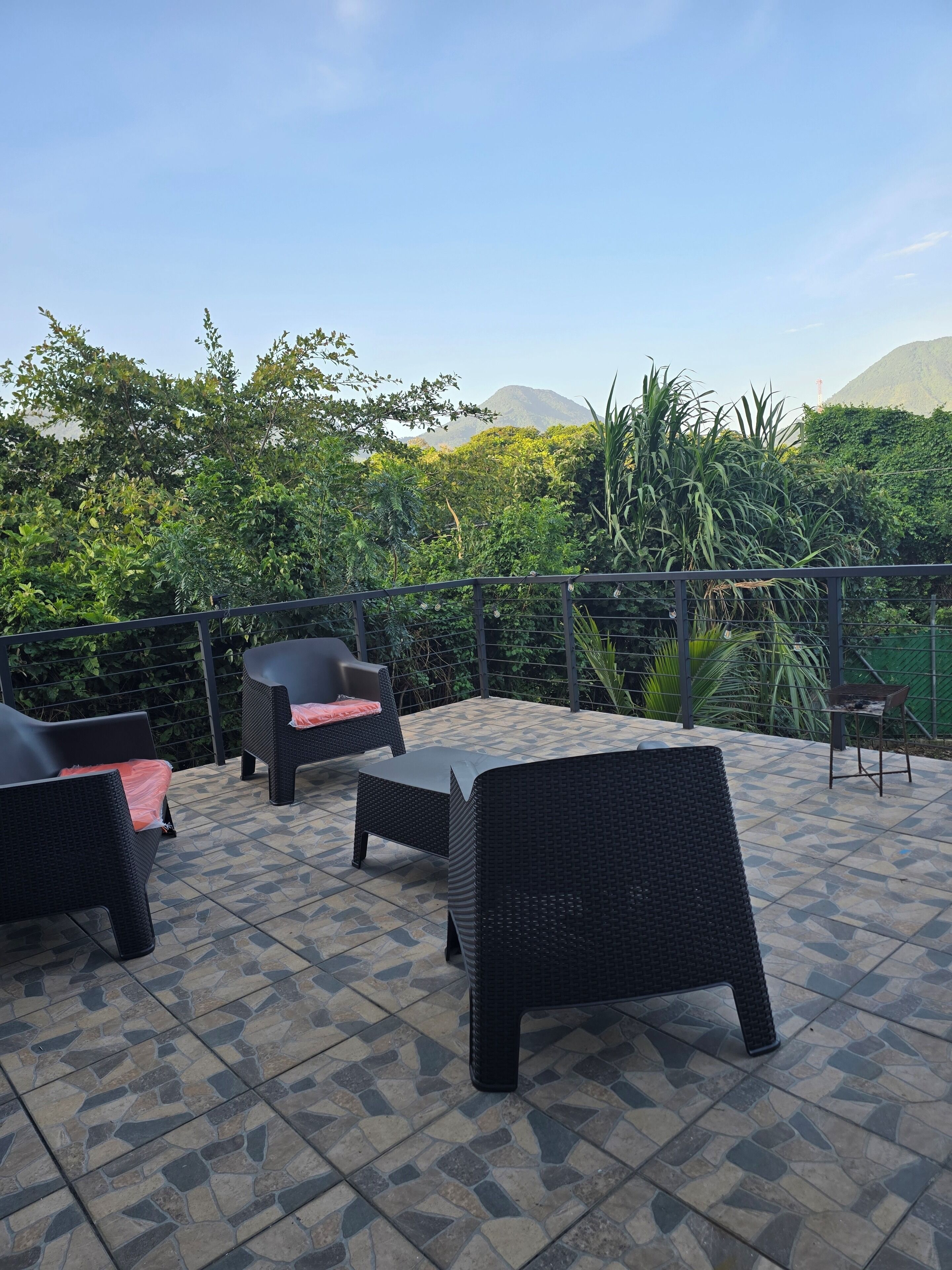 Een terras buiten met rieten meubilair en een betegelde vloer, omringd door weelderige begroeiing en een berg op de achtergrond.