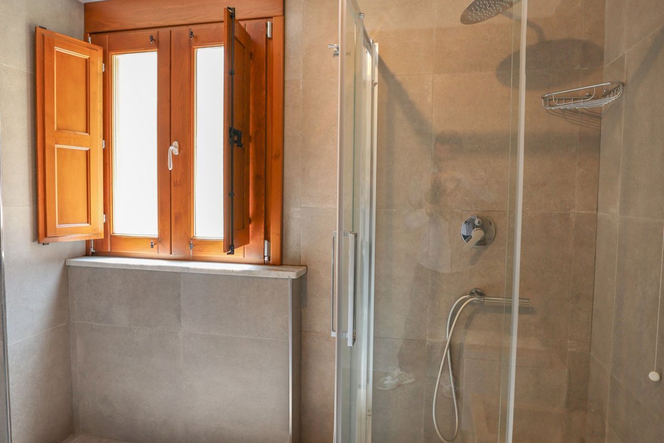 Ein Badezimmer mit Duschkabine aus Glas, einem Holzfenster mit geöffneten Fensterläden und einem gefliesten Boden.