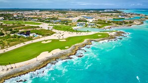 Vue aérienne d’un complexe hôtelier de bord de mer comprenant un terrain de golf avec plusieurs verts, une plage de sable et une côte rocheuse.