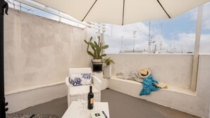 Un balcon avec un canapé blanc, une petite table sur laquelle se trouvent une bouteille et un livre, une plante en pot et un chapeau posé sur le rebord.