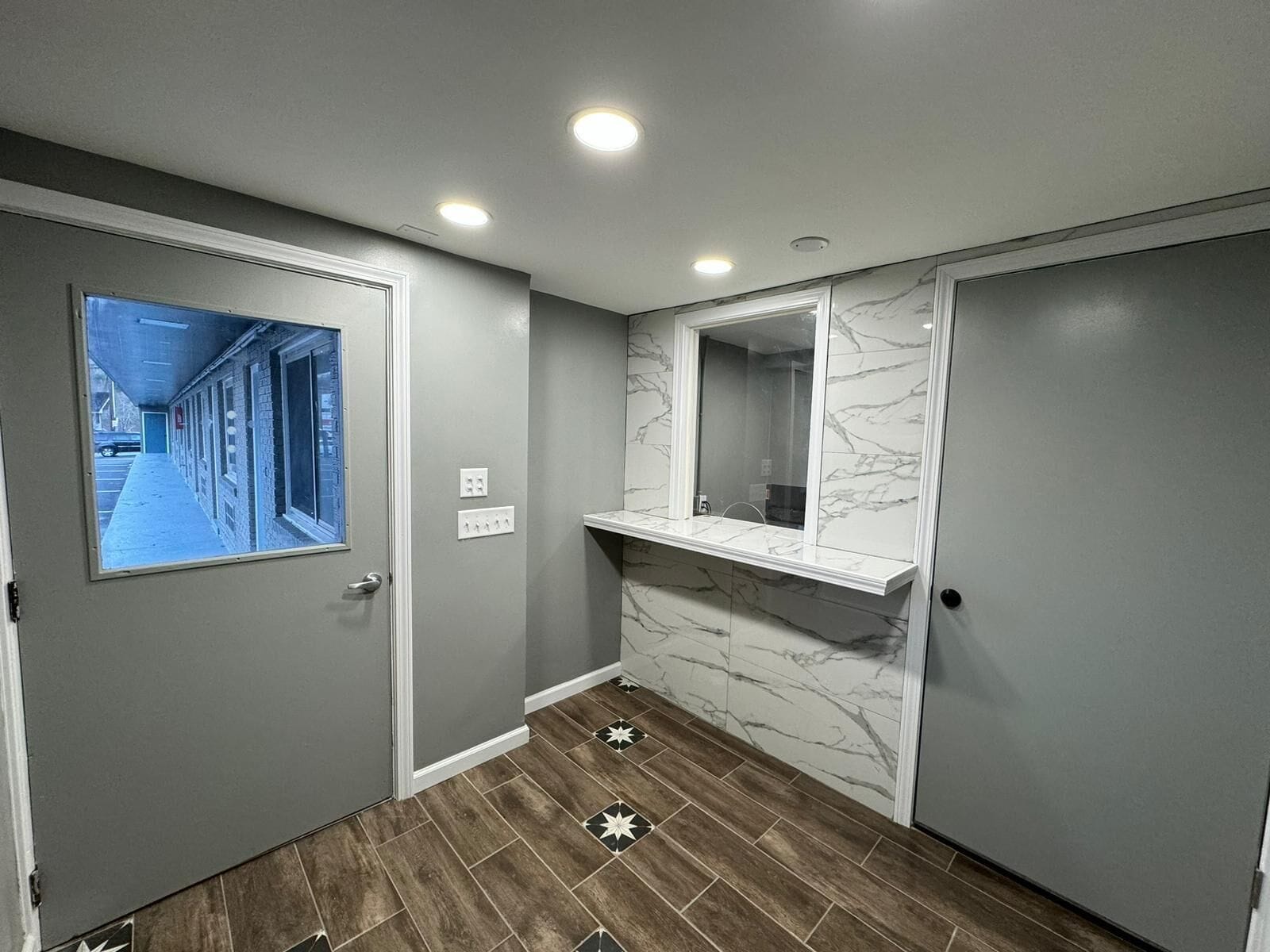 A hallway with a door leading to a parking area, a marble wall with a mirror, and a tiled floor with a star pattern.