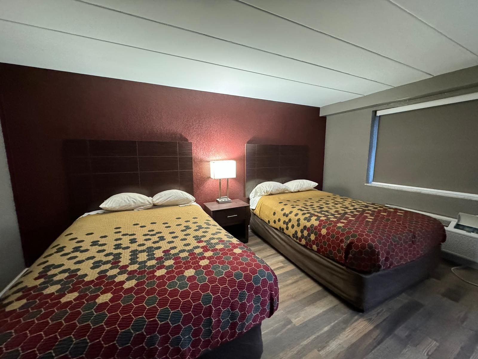 A hotel room with two beds, each with a patterned bedspread, a nightstand with a lamp, and a window with a view.