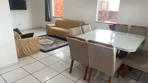 A living room with a sofa, a dining table, and chairs. There is a TV mounted on the wall.