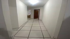 A narrow hallway with white tiled floors and walls, leading to a door at the end.