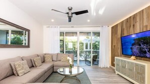 Ein modernes Wohnzimmer mit einem Ecksofa, einem runden Couchtisch, einem Deckenventilator, einem Fernseher und einer Glastür zum Außenbereich.
