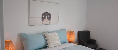 A neatly arranged bedroom with a bed, pillows, a chair, and a wall-mounted picture.