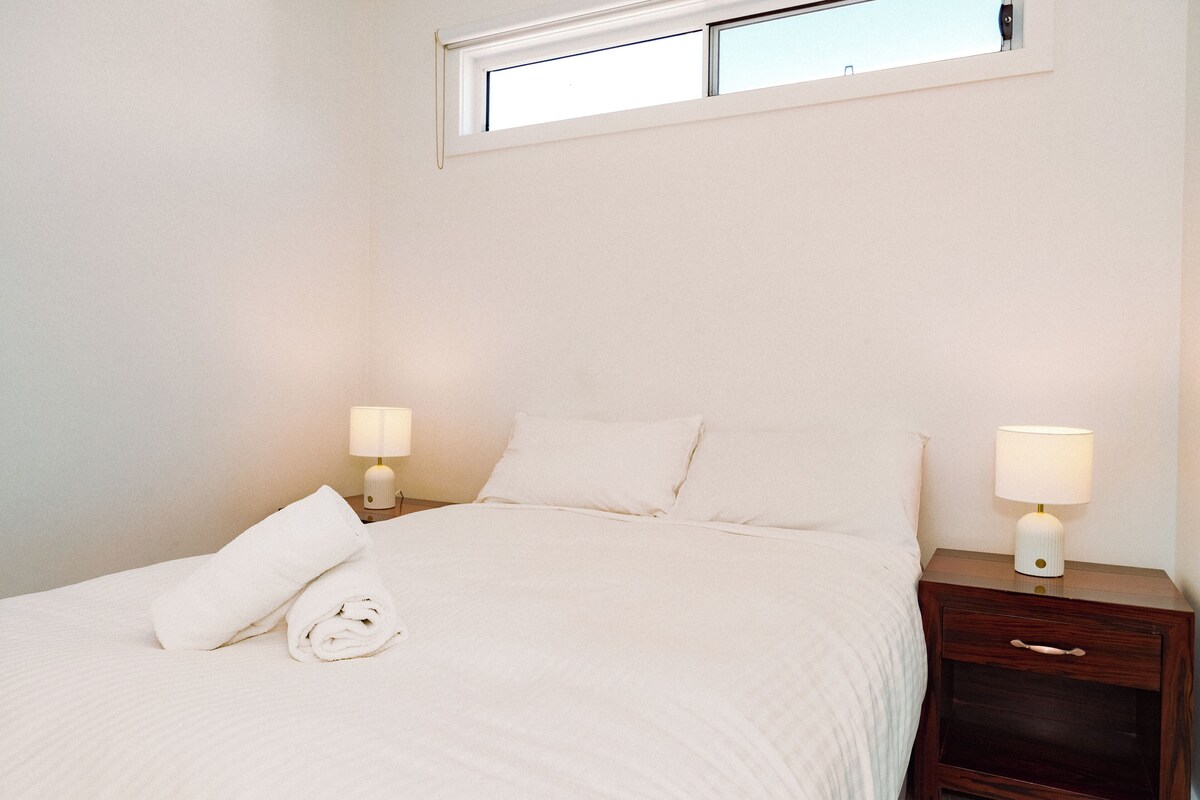 A neatly made bed with a rolled white towel, two bedside lamps, and a small wooden nightstand with a drawer.