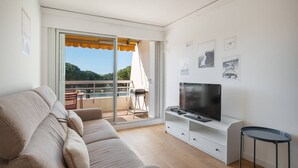 A modern living room with a sofa, a flat-screen TV on a white stand, and a balcony with a view of the sea.