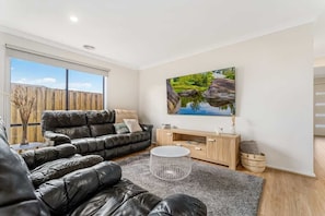 A modern living room with a black leather sofa, a round coffee table, a wooden TV stand, and a large window.