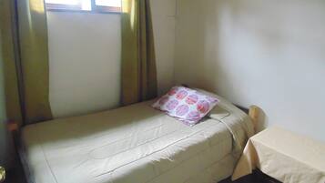 Quarto pequeno com uma cama de solteiro, um lençol, uma almofada, uma mesa com toalha e uma janela com cortinas.